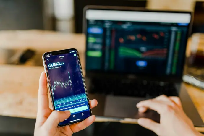 Person holding smartphone with trading app in front of laptop displaying financial charts